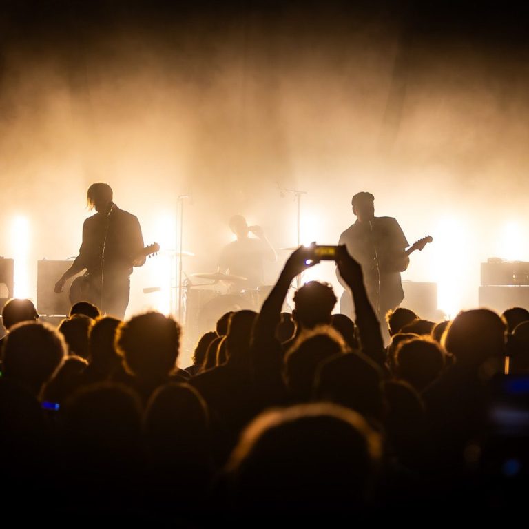 Silhouetted band on stage with bright lights and crowd filming with phones.